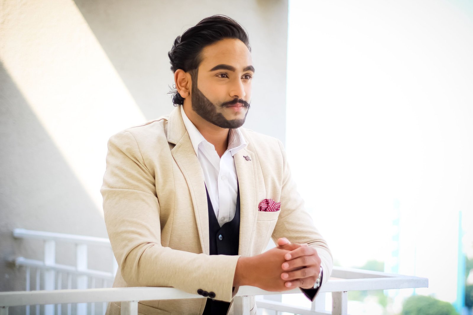 A poised man in a beige suit stands on a balcony, exuding elegance and sophistication.